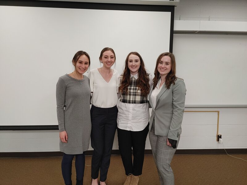 Four college-age women standing together in front of a projector screen. 