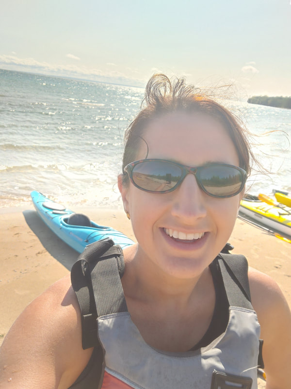 Vanessa standing on Lake Michigan shoreline; blue and yellow kayaks in background. 